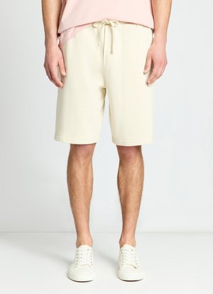 Uomo che indossa pantaloncini crema con coulisse, t-shirt rosa chiaro e scarpe da ginnastica bianche, in piedi su uno sfondo chiaro e semplice.