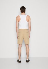 Bermudas beige con bolsillos laterales, hechas de una tela ligera. Combinadas con una camiseta sin mangas blanca y zapatillas negras, vistas desde la parte de atrás.