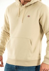 Felpa beige realizzata in tessuto misto cotone, dotata di una tasca frontale, polsini e orlo ribattuti, e una piccola etichetta logo sul petto.