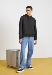 Sudadera con capucha negra, con un bolsillo frontal y un pequeño logo, combinada con jeans de mezclilla azul claro y zapatillas negras sobre un suelo amarillo.