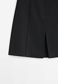 Jupe noire mi-longue avec une fente centrale à l'avant et des détails subtils de couture, posée à plat sur une surface blanche.