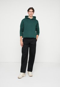 Νεαρός άνδρας στέκει, φορώντας σκούρο πράσινο φούτερ, μαύρο παντελόνι και λευκά αθλητικά παπούτσια, με τα χέρια στις τσέπες, ουδέτερη έκφραση, απλό λευκό φόντο.