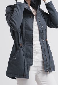Parka de color gris oscuro con cremallera frontal, capucha ajustable y dos bolsillos laterales con detalles de botones. La tela tiene una textura suave y ligera.