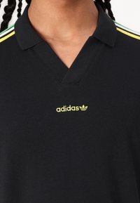 Czarna koszulka polo z dekoltem w serek. Posiada żółte logo Adidas i trzy poziome paski w kolorach zielonym, żółtym i czarnym na ramionach.