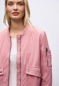 Giovane donna che indossa una giacca bomber rosa a costine sopra una maglietta bianca, con tasca con zip sulla manica, su uno sfondo chiaro e uniforme.