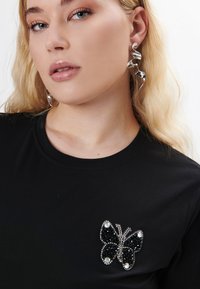 Sort t-shirt med et dekorativt sommerfugle-applikation lavet af sorte paljetter og sølvdetaljer, placeret mod en glat stofbaggrund.