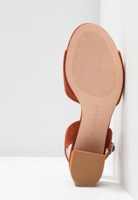 Sandale femme en daim marron avec talon bloc, semelle texturée et bride à boucle, vue de dessous sur fond blanc.