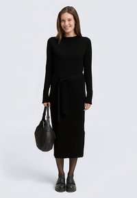 Schwarzes langärmliges Kleid mit gerader Silhouette, geschnürter Taillendetail und weicher Textur. Kombiniert mit einer schwarzen Schultertasche und klobigen Schuhen.