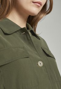 Camicia verde oliva con colletto bottonato, due tasche sul petto e una texture liscia. Presenta un singolo bottone al collo e dettagli di cucitura.