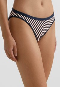 Nő tengerészkék és fehér csíkos bikini alsót visel, amelyen átlós mintázat és egyszínű tengerészkék szegély található, világos háttér előtt.