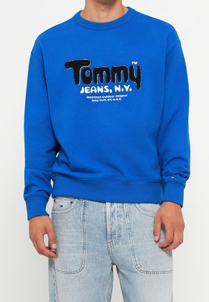 Man draagt een felblauwe trui met de tekst "Tommy Jeans, N.Y." en lichtblauwe spijkerbroek, staande tegen een effen achtergrond.