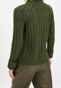 Grüner gestrickter Pullover mit geripptem Textur, hohem Kragen und umgeschlagenen Manschetten. Kombiniert mit braunen engen Hosen, die ein lässiges Design zeigen.