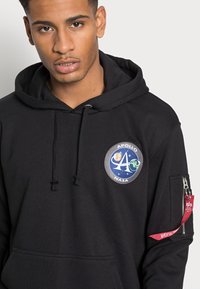Sweatshirt preto com um patch redondo "Apollo NASA", capuz com cordão, bolso frontal e bolso com zíper na manga com uma aba vermelha.