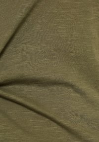 Tissu vert olive avec une légère texture ; matériau doux et extensible présentant un motif de tissage subtil et inégal. La lumière se reflète doucement.