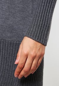 Graue gerippte Strickpulloverärmel mit einem strukturierten Muster und eng sitzendem Handgelenk; Hand ruht auf dem Stoff und zeigt das engmaschige Strickmuster und die Weichheit.