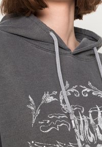 Osoba nosząca szaro-sweterek z kapturem z białym wzorem kwiatowym i widocznymi sznureczkami w okolicy kołnierza.