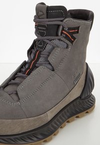 Botte de randonnée imperméable grise avec dessus en suède et mesh, équipée de ferrures noires, semelle en caoutchouc, accents contrastants orange et design texturé.