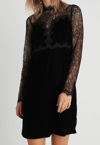 Robe noire avec un corsage en velours texturé et des manches longues en dentelle. Présente un design en dentelle festonné au niveau de la poitrine et une coupe décontractée.