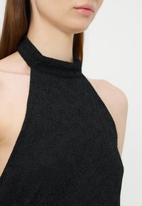 Femme portant un haut noir texturé à col halter, montrant son cou, ses épaules et une partie de son visage sur un fond clair uni.