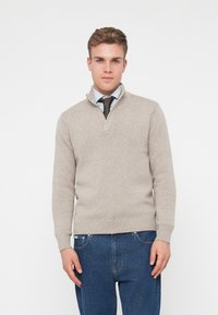 Jasno beżowy sweter dzianinowy z kołnierzem na ćwierć suwaka, ściągaczami na rękawach i u dołu. Nosi się go na niebieskiej koszuli oraz w połączeniu z dżinsami w kolorze niebieskim.