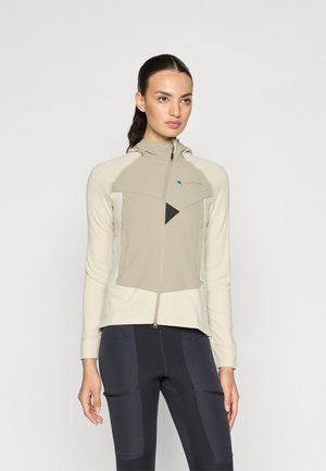 Capuchonjack in lichtbeige en tan met een gestructureerde stof; heeft een rits aan de voorkant, schuine kleurvlakken en logodetail. Gecombineerd met een donkere legging.