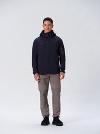 Veste imperméable navy avec poches zippées, capuche ajustable et accents de zipper jaune contrastants, associée à un pantalon beige zippé et des baskets noires.