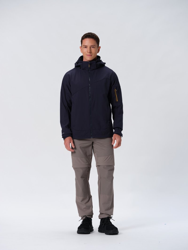 Veste imperméable navy avec poches zippées, capuche ajustable et accents de zipper jaune contrastants, associée à un pantalon beige zippé et des baskets noires.