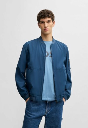 Giacca bomber blu con una superficie liscia, colletto e polsini a costine, e cerniera frontale. Indossata sopra una camicia blu chiaro e abbinata a denim.