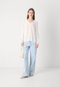 Lichte, crèmekleurige blouse met een v-hals en koordje, gecombineerd met lichtblauwe wijde jeans. Het model houdt een beige handtas vast en draagt naak kleurige pumps.
