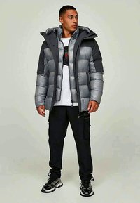 Homem a usar um casaco acolchoado em dois tons de cinza, um sweatshirt gráfico branco, calças cargo pretas e ténis preto e branco, de pé com as mãos relaxadas.