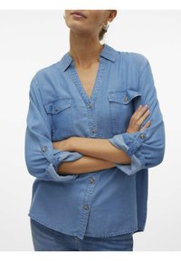 Blusa in denim azzurro chiaro con scollatura a V, maniche lunghe arrotolate, due tasche sul petto e dettagli con bottoni. Texture morbida, design casual.