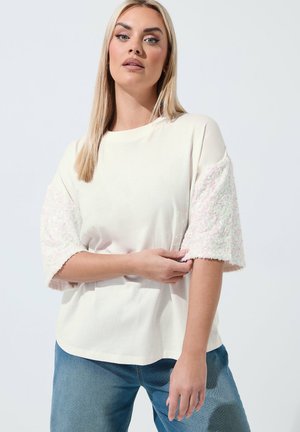 Femme aux cheveux blonds raides portant un haut blanc ample avec des manches courtes texturées et un jean bleu, posant avec un bras croisé sur son corps.