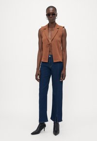 Colete castanho sem mangas com um nó à frente, combinado com jeans de perna reta azul-escuro com fendas laterais e saltos agudos pretos.