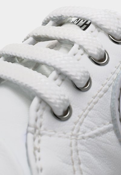 Κοντινή λήψη λευκών κορδονιών αθλητικών παπουτσιών διαπερασμένων από μεταλλικά οπές σε υφασμένο δερμάτινο επάνω μέρος κοντά σε ραμμένη ετικέτα λογότυπου.