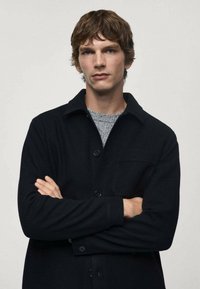 Giacca camicia nera con bottoni, realizzata in tessuto morbido, dotata di taschino sul petto, maniche lunghe e colletto.