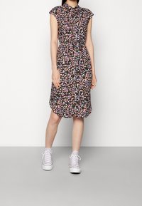 Robe noire à fleurs avec des manches courtes, taille élastique et devant à boutons ; petites fleurs colorées ; associée à des baskets roses clair.