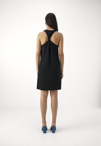 Nainen seisoo selin, päällään hihaton musta mekko, jossa on kierretty selkätalous, sekä siniset paksupohjaiset sandaalit valkoisella taustalla.