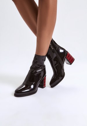 Botas de tobillo de charol negro con un acabado brillante, punta cuadrada y un tacón bloque de cuadros rojos chunky. Cierre de cremallera en el lateral para facilitar su uso.