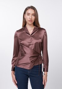 Πουκάμισο τύπου σατέν σε πλούσιο καφέ χρώμα με γιακά, μακριά μανίκια, κουμπιά και λεπτομέρεια δεσίματος στο πλάι. Συνδυασμένο με σκουρόχρωμο μπλε τζιν.