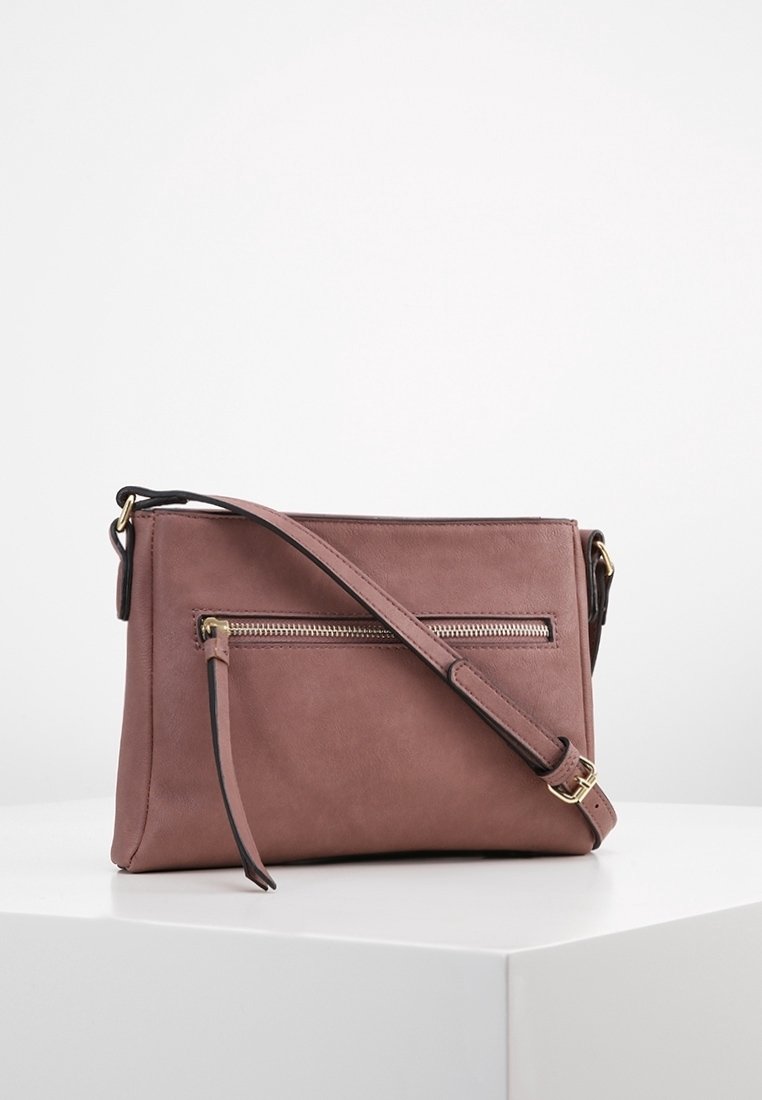 Bolso cruzado rectangular de cuero malva con bolsillo frontal con cremallera, correa ajustable y herrajes en tono dorado sobre un pedestal blanco.