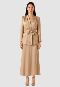 Blusa beige stile avvolgente con maniche lunghe, legata in vita, abbinata a una gonna lunga coordinata fino al pavimento. Tessuto liscio e strutturato.