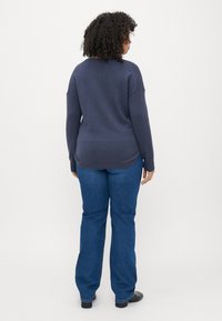 Mornarska pulover z rebrastim robom, v paru z modrimi razširjenimi jeans hlačami. Pulover ima sproščen kroj in teksturirane rokave. Vidni so črni čevlji.