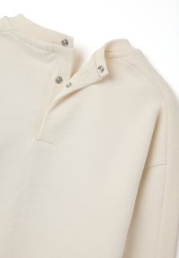 Felpa color beige chiaro con colletto a costine e patta con bottoni; tessuto morbido con una superficie liscia; presenta due bottoni a pressione argentati.