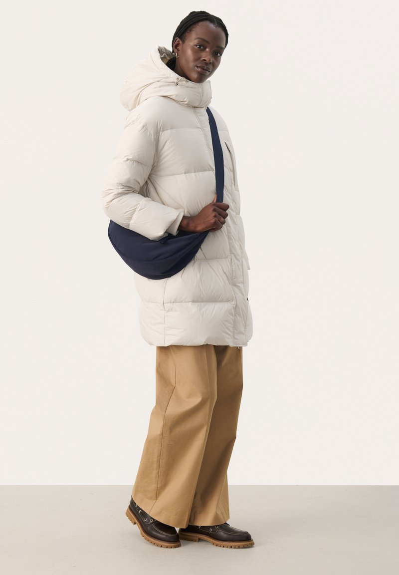 Puchowa kurtka parka z kapturem, z zamkiem błyskawicznym z przodu i kieszeniami. W połączeniu z jasnobrązowymi spodniami o szerokich nogawkach i ciemnobrązowymi botkami.