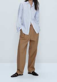 Ljusblå och vit randig skjorta med knäppning, i en avslappnad passform, parat med lösa bruna byxor och svarta slip-in sandaler.