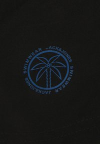 Tissu noir avec un logo circulaire bleu représentant un palmier, entouré par le texte "JACK&JONES MAILLOTS DE BAIN." Texture douce, design décontracté.