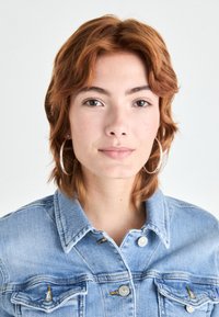 Svijetloplava traper jakna s kopčanjem na gumbe i dva prednja džepa. Model ima srednje dugačku valovitu crvenu kosu i velike srebrne okrugle naušnice.