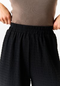 Black textured wide-leg pants featuring an elastic waistband, paired with a fitted brown top. Focus on fabric texture and design details.