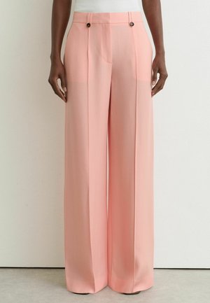 Lyserøde bukser med høj talje og vide ben, for-plieringer og knappedetaljer, båret med en hvid top mod en ensfarvet baggrund.