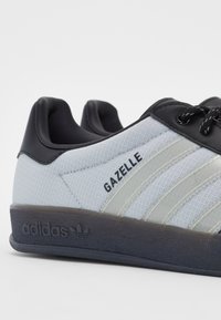 Bílá a černá teniska Adidas Gazelle zobrazující bok s texturovanou látkou, černým jazykem na patě, tkaničkami a tmavou podrážkou s logem Adidas.